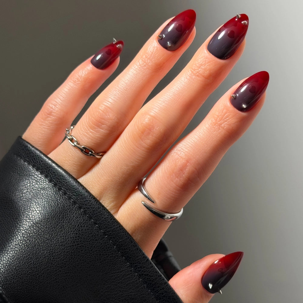 A hand with short almond nails in glamnetic’s “Lil Devils” design, featuring a glossy red-to-black ombre, accessorized with silver rings and a black leather sleeve.