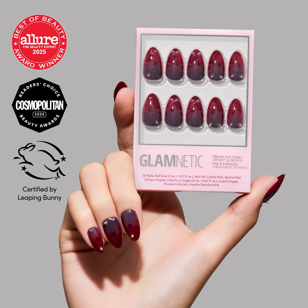 A hand with glossy, burgundy ombre press-on nails holds a box of Lil Devils red jewelry nails from glamnetic. Award logos and a cruelty-free certification appear on a gray background, accentuating these stylish short almond-shaped nails. - Product image