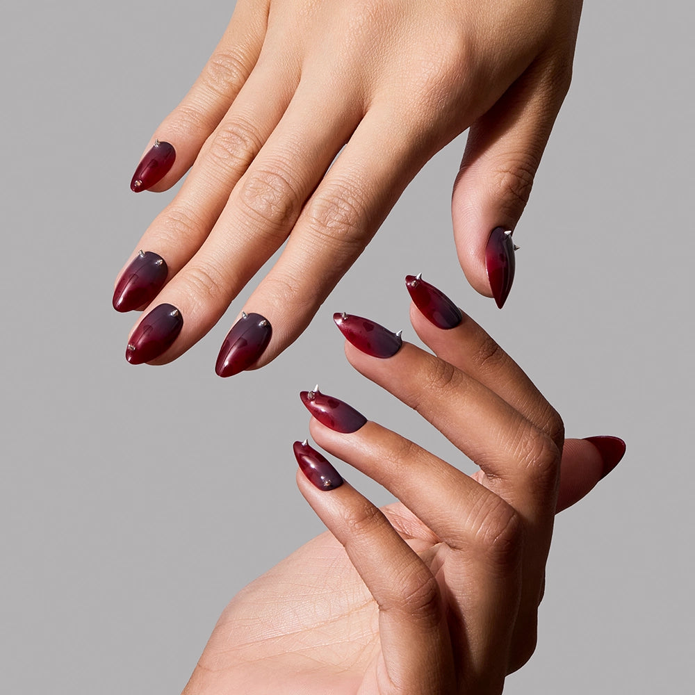Two hands with short almond nails are painted in a glossy, dark red and black ombré design—evoking the look of red jewelry nails—featuring the Lil Devils press-ons by glamnetic, displayed against a plain gray background. - Product image