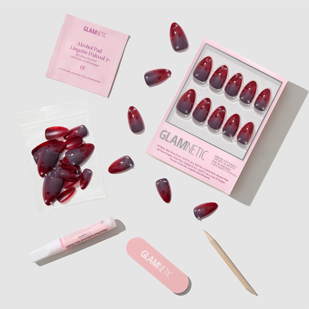 Introducing Lil Devils by glamnetic—a set of short almond press-on nails in vibrant red and purple shades, adorned with red jewel nail accents. The set includes an alcohol pad, nail glue, a mini nail file, a wooden cuticle stick, and extra nails—all elegantly arranged against a light background. - Product image