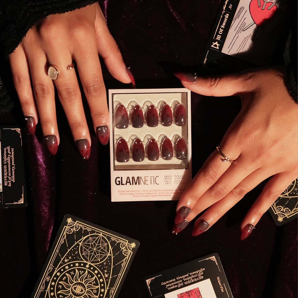 Two hands display red and black ombré Lil Devils press-on nails in a chic short almond shape next to a glamnetic nail set box, tarot cards, and astrological designs on a dark purple surface. - Product image