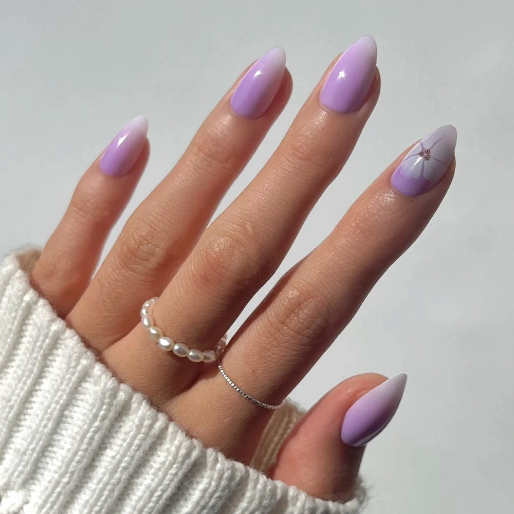 A hand with long, almond-shaped glamnetic Lilac Reef nails in an ombre design, featuring a floral accent on one nail. The person wears pearl and silver rings, and a white knitted sleeve peeks into the frame.