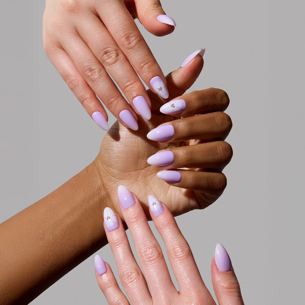 Three pairs of hands showcase glamnetic’s Lilac Reef press-on nails, featuring almond-shaped lavender ombré designs—some adorned with tiny flowers—artfully overlapped against a plain light gray background. - Product image