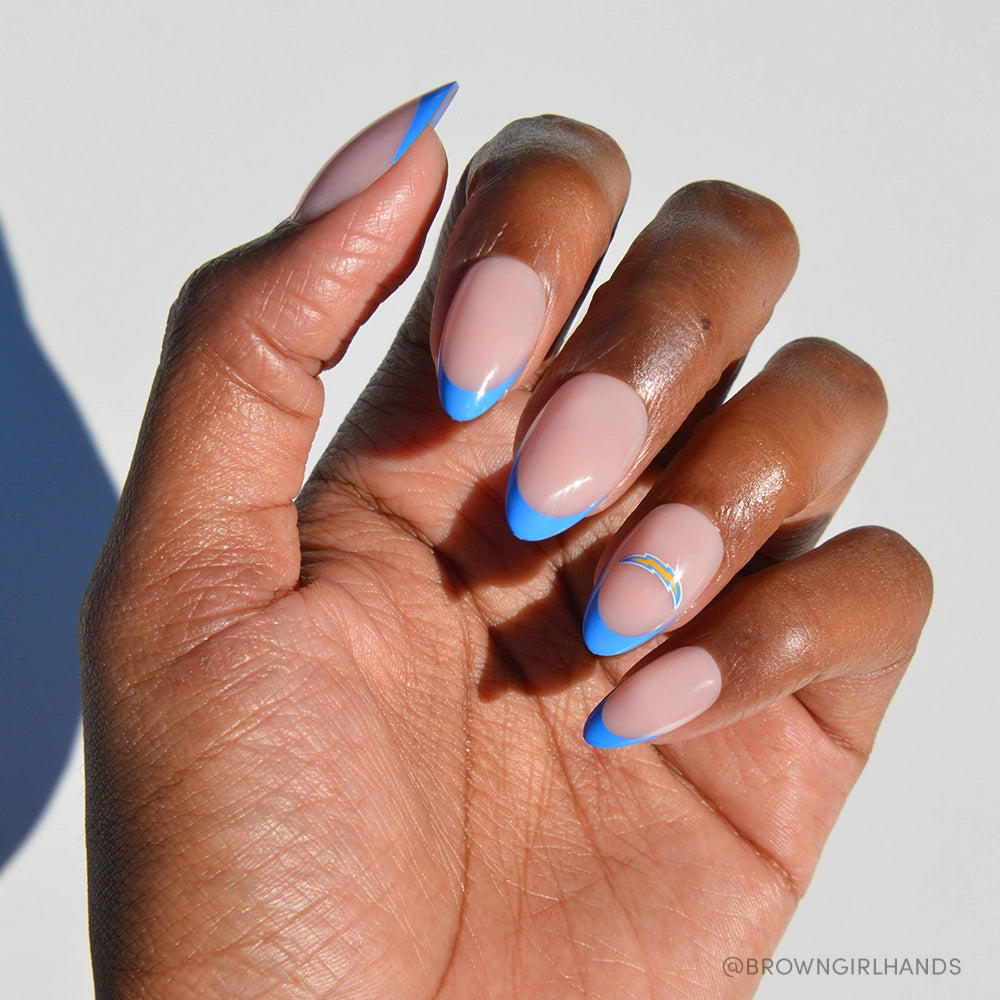 A deep brown hand wears glamnetic&#39;s Los Angeles Chargers Press-On Nail Set: almond-shaped with a nude base, bright blue French tips, and a rainbow accent nail, all set against a white background.