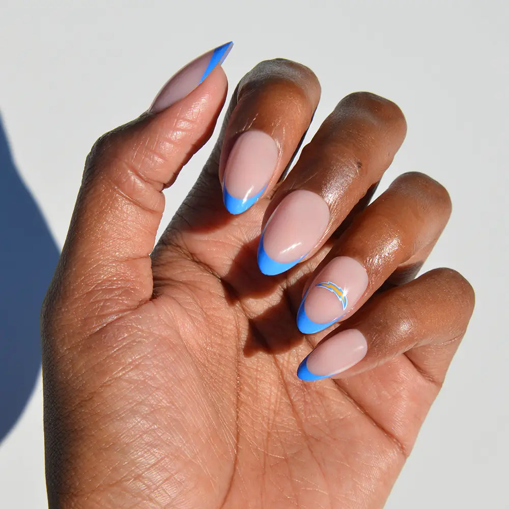 A hand with almond-shaped NFL Press-On Nails by glamnetic features a nude base, bright blue French tips, and a small rainbow design on the ring finger. - Product image