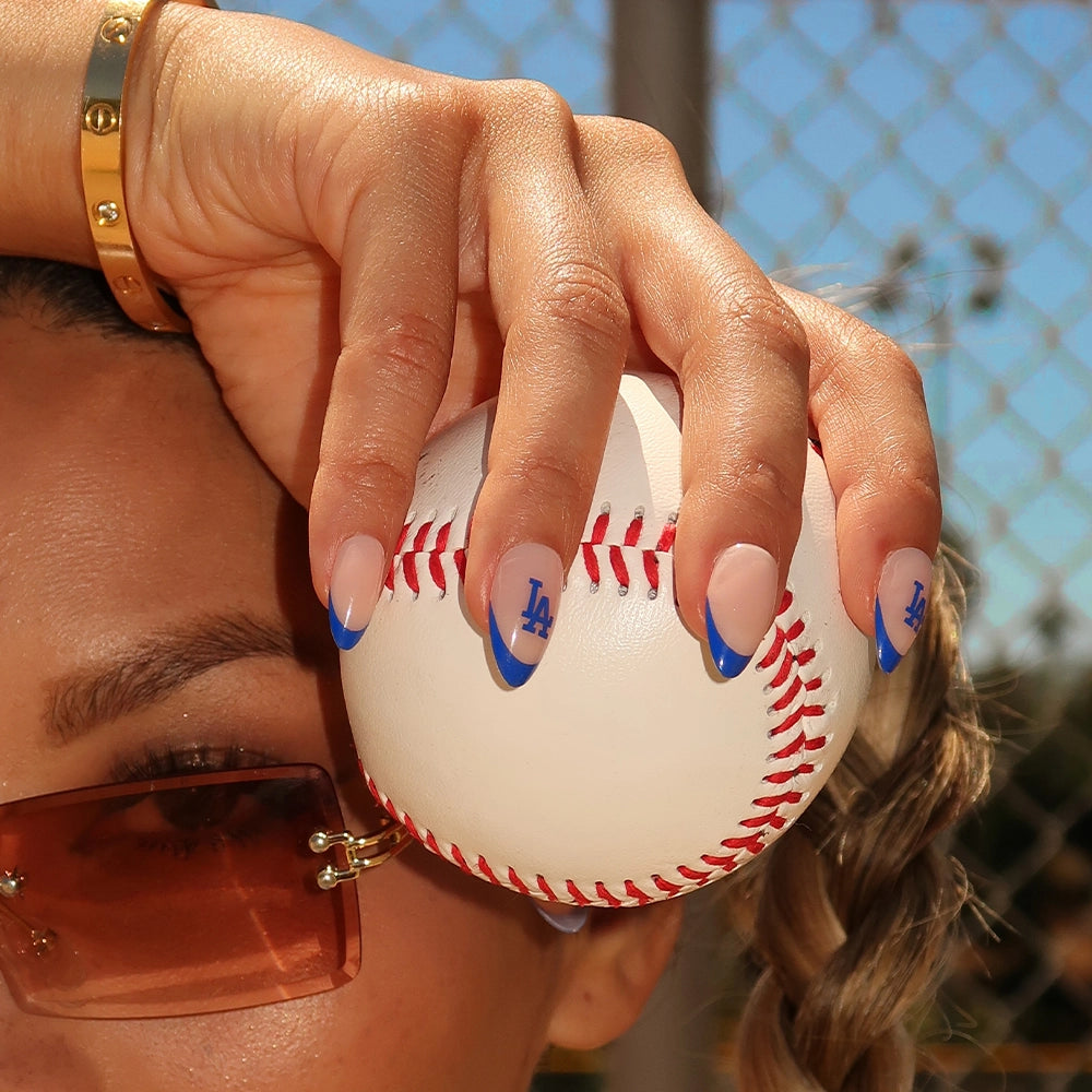 A person holds a baseball, displaying manicured nails with blue tips and LA logos from the glamnetic Los Angeles Dodgers Press-On Nail Set, while wearing sunglasses and a gold bracelet by a chain-link fence. - Product image
