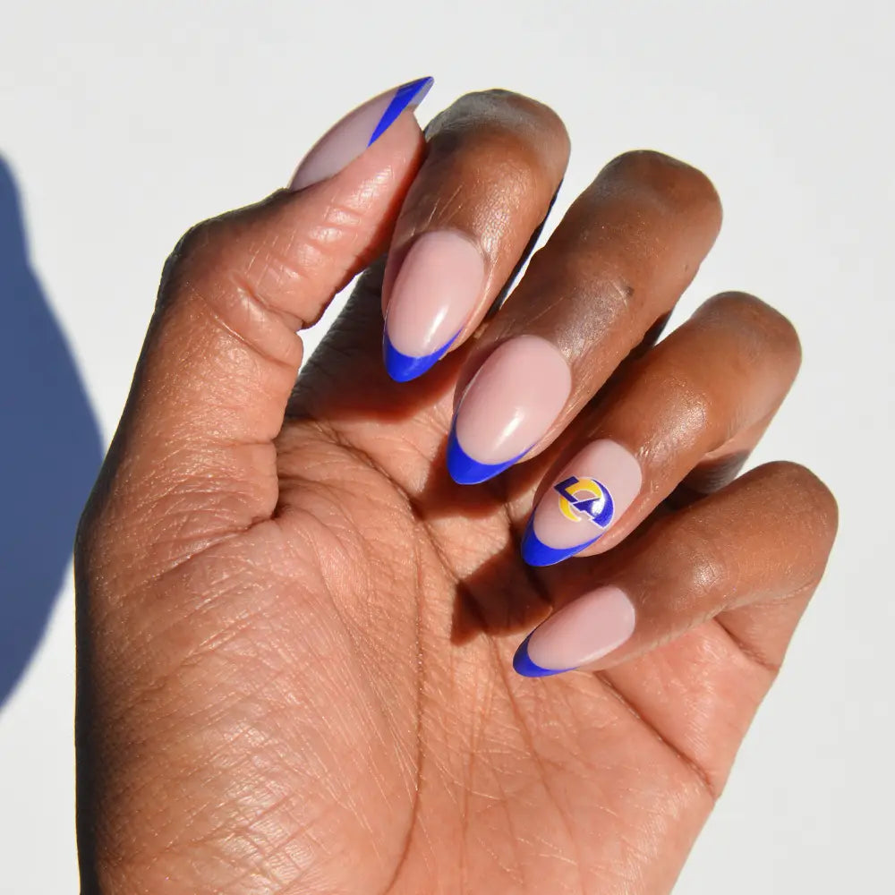A hand wearing glamnetic NFL Press-On Nails, featuring almond-shaped nails with nude polish, blue French tips, and a Los Angeles Rams logo on the ring finger. - Product image