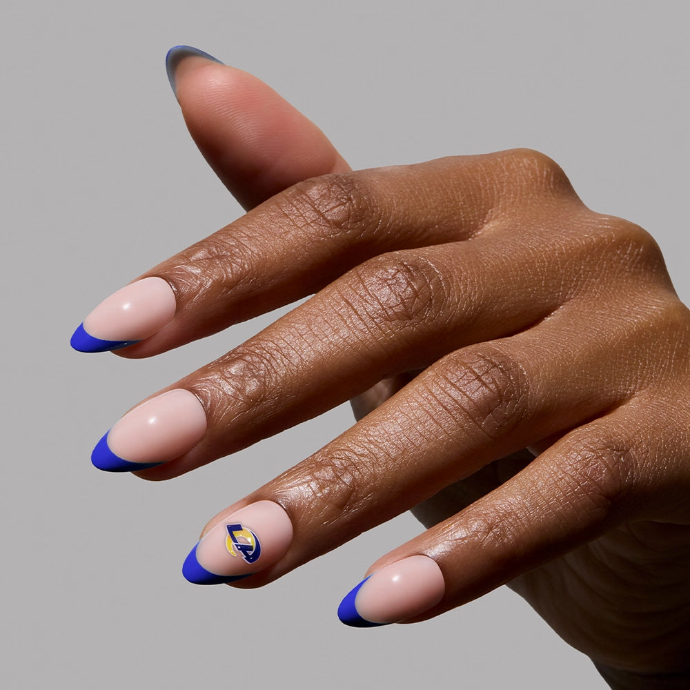 A hand wears the glamnetic Los Angeles Rams Press-On Nail Set, with almond-shaped nude nails and bright blue French tips. The ring finger features a small gold and blue accent. The background is plain gray. - Product image