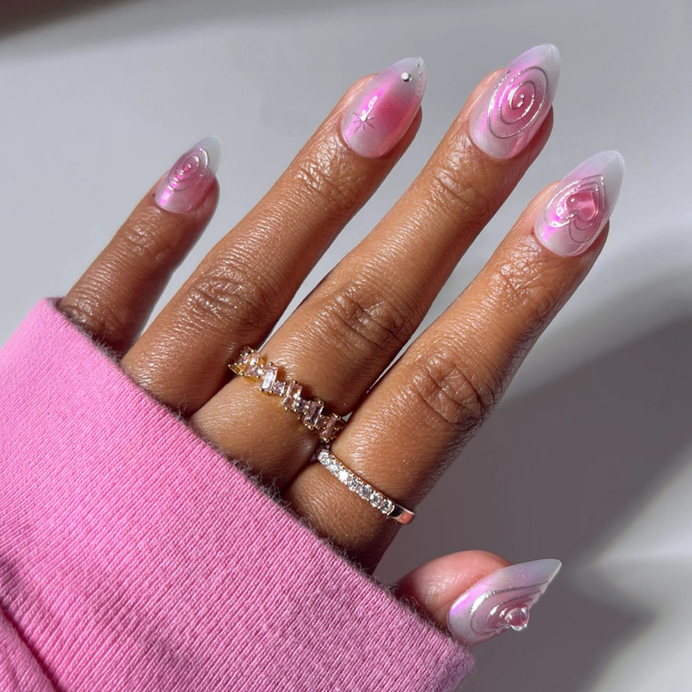 A hand displays glamnetic’s Love Current press-on nails in a short almond shape, adorned with pink and white swirl nail art and accented by delicate rhinestones. Gold and diamond rings embellish the fingers, while a pink sleeve partially drapes the hand, drawing attention to the chic elegance of these Love Current nails. - Product image