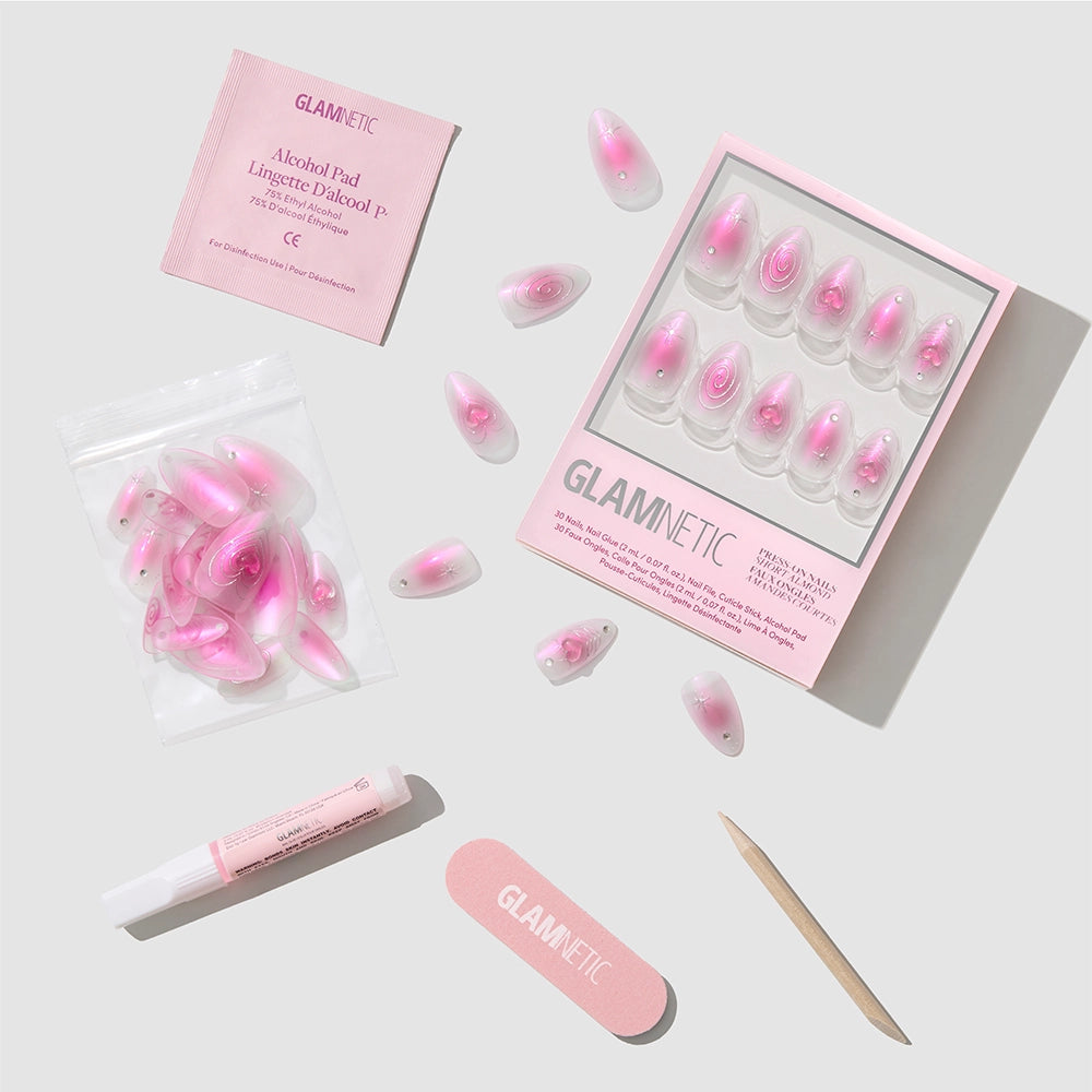 The Love Current press-on nails by glamnetic, featuring a pink marble design in a short almond shape, are displayed on a light grey background along with glue, a wooden cuticle stick, nail file, alcohol pad, and extra nails. - Product image