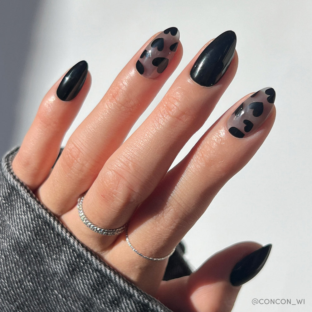 A hand with long almond Love Me Not nails by glamnetic shows glossy black tips and two accent nails with sheer bases and black heart designs, paired with thin silver rings and a gray sleeve.