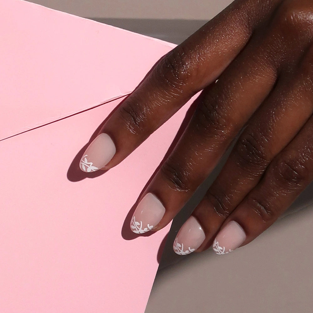 A hand with short, almond-shaped nails painted in glamnetic’s Love Preserved, a pale nude shade with delicate white leaf tip design, rests on a pale pink envelope. - Product image