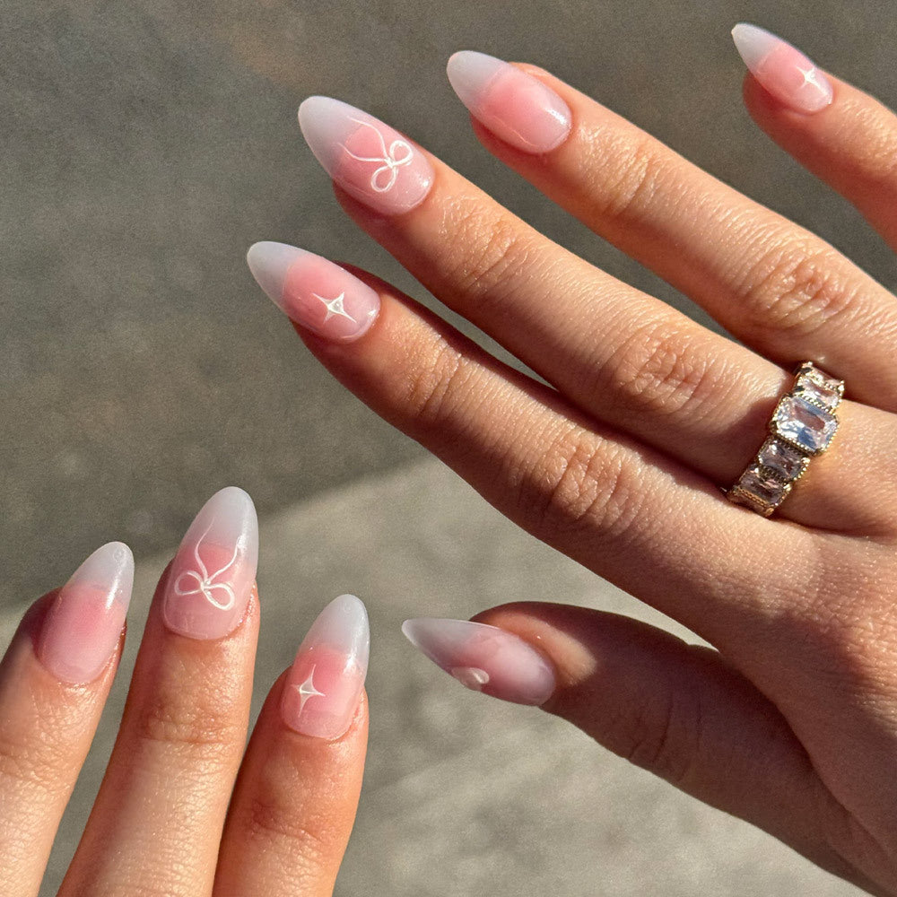 A close-up of hands with Glamnetic Lovers Knot semi-transparent pink nails, almond-shaped with a pink-to-white ombré and delicate white line art. One hand features a gold ring with rectangular clear stones.