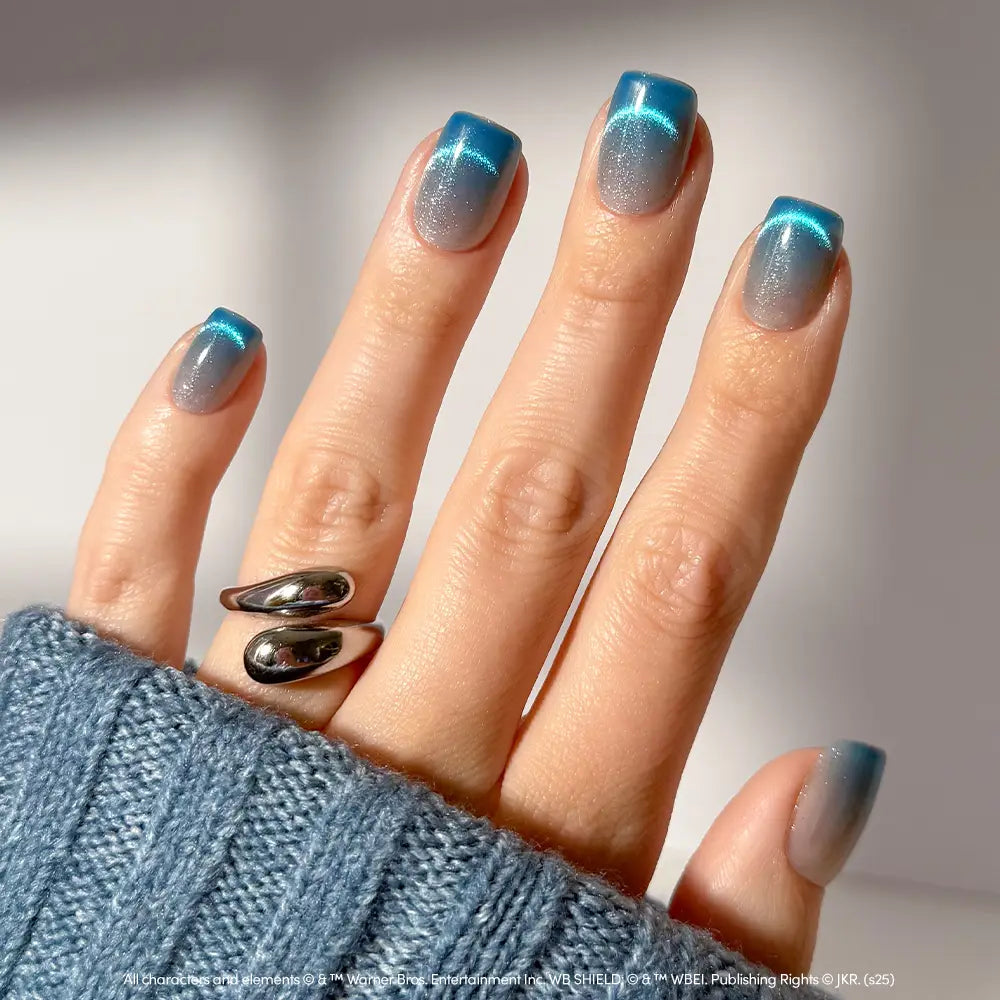 A hand displays glamnetic’s Lumos velvet blue gradient nails with a blue-to-clear ombre effect, complemented by a chunky silver ring and a blue sweater sleeve partially covering the fingers.