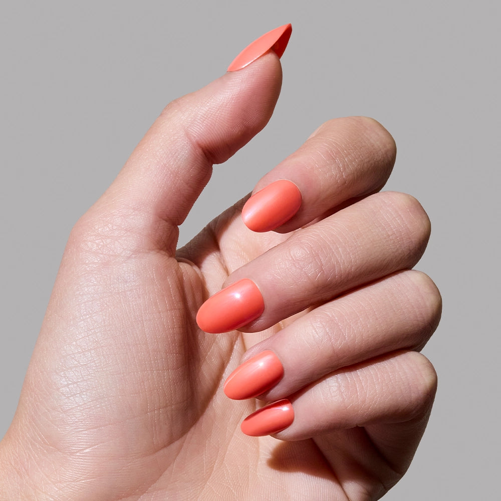 A hand with medium-length oval nails painted in glamnetic’s Lush Coral, a smooth, glossy vibrant coral polish, against a plain gray background. - Product image