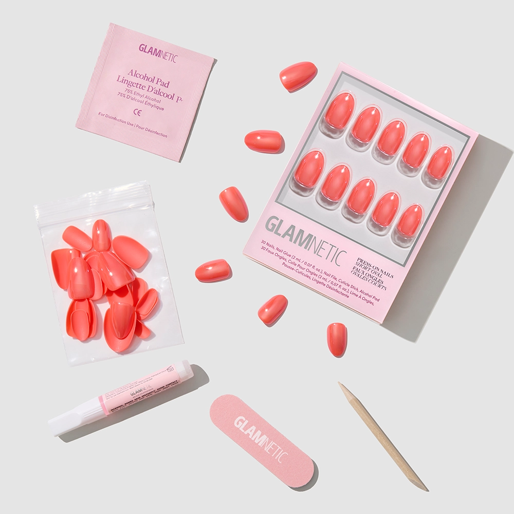 The Lush Coral set by glamnetic includes vibrant coral short oval nails, extra nails, nail glue, a wooden cuticle stick, an alcohol pad, and a nail file—all arranged neatly on a light surface with the glamnetic logo visible. - Product image