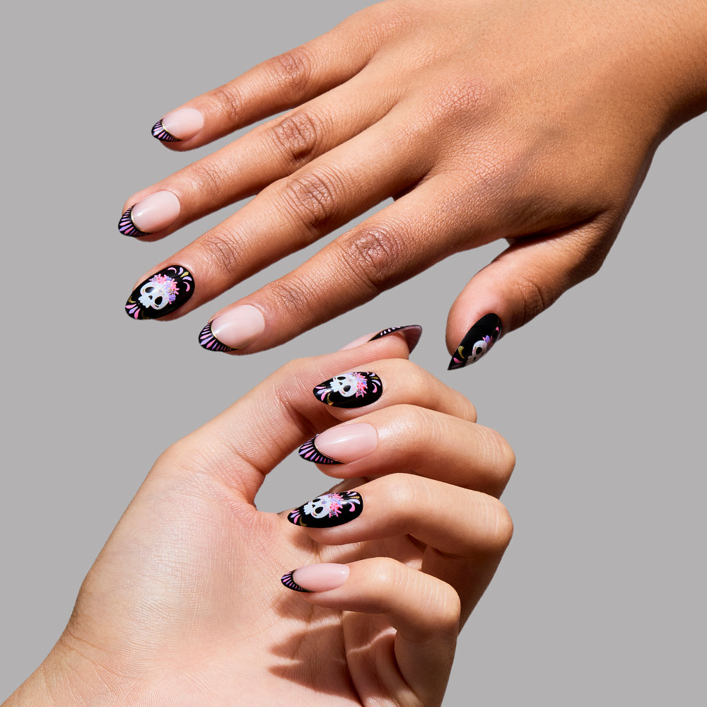 Two hands with short almond nails wearing glamnetic’s Mani De Los Muertos, featuring a black base, white floral accents, and striped tips inspired by Day of the Dead designs. The background is plain gray. - Product image