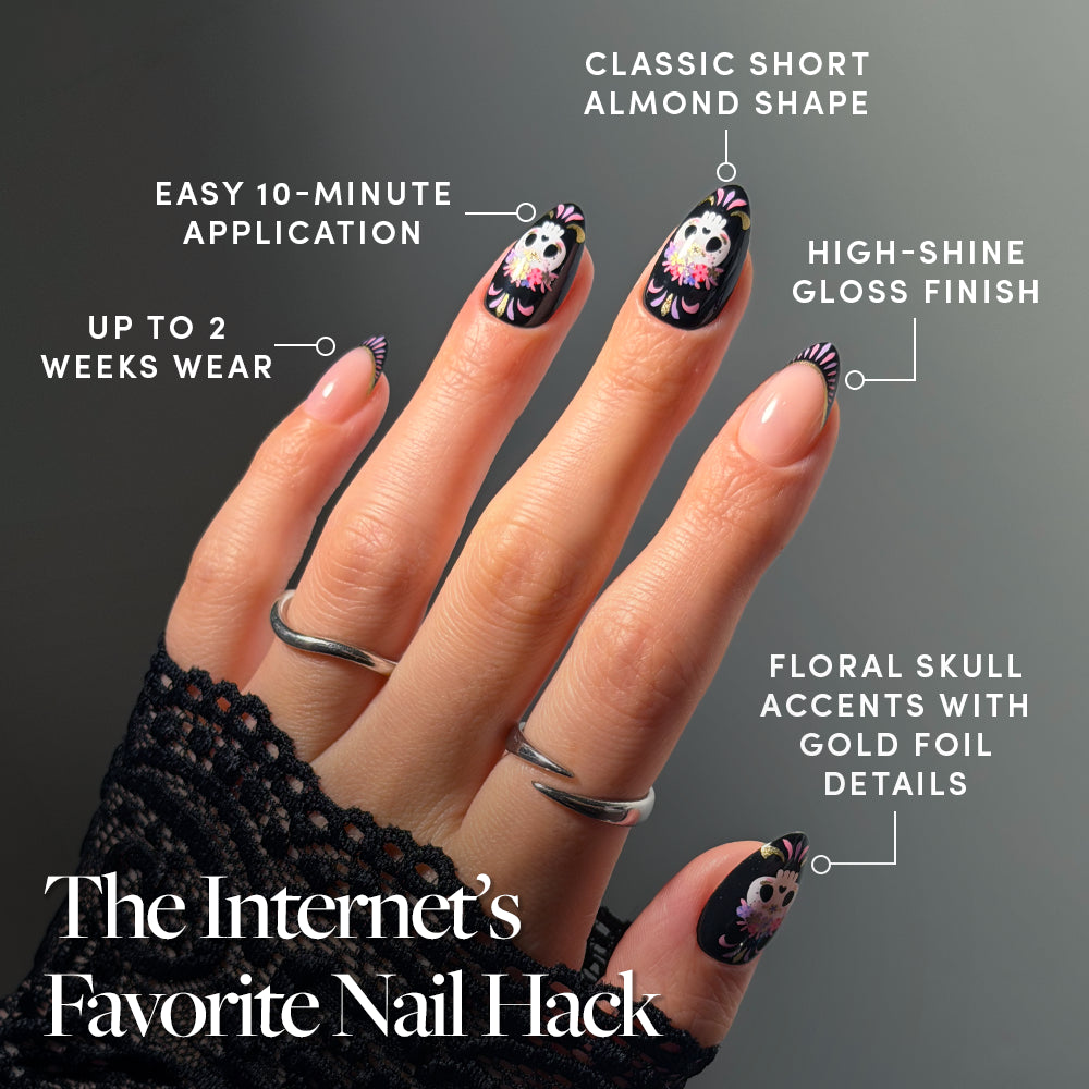 A hand adorned with glamnetic’s Mani De Los Muertos press-on nails showcases black floral skull nail art inspired by Day of the Dead, featuring intricate gold details on a glossy almond-shaped set. The look highlights an easy 10-minute application, durable two-week wear, and a high-shine finish. The hand is accessorized with silver rings and a black lace sleeve. - Product image