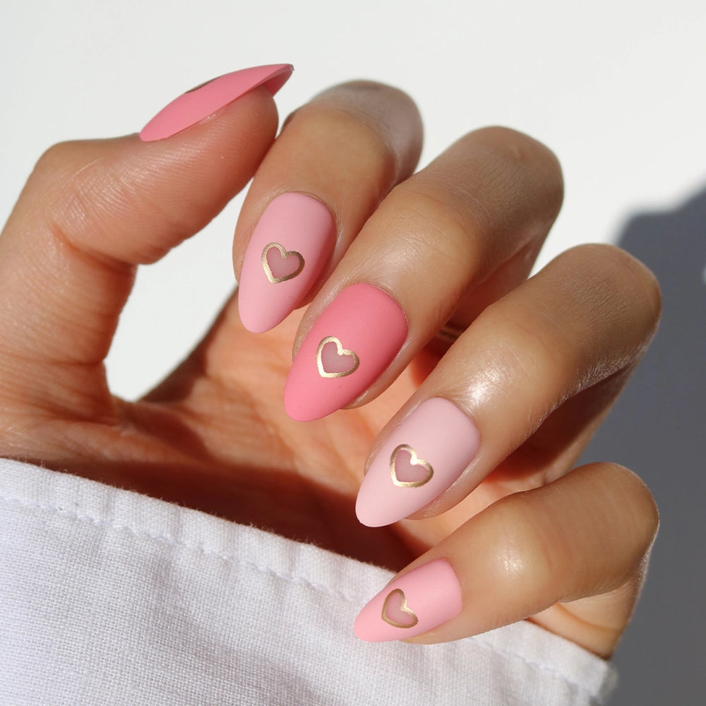 A hand wears glamnetic’s Matte in Love short almond press on nails in soft pink matte, each adorned with a dainty gold heart cutout, perfectly complementing the white sleeve.
