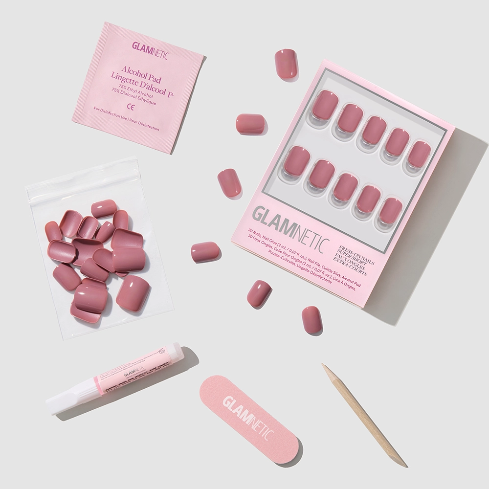 A flat lay of the glamnetic Mauve Cutie press-on nails kit featuring glossy mauve round nails, extra nails, an alcohol pad, nail file, cuticle stick, nail glue, and a small resealable pouch on a light grey background. - Product image