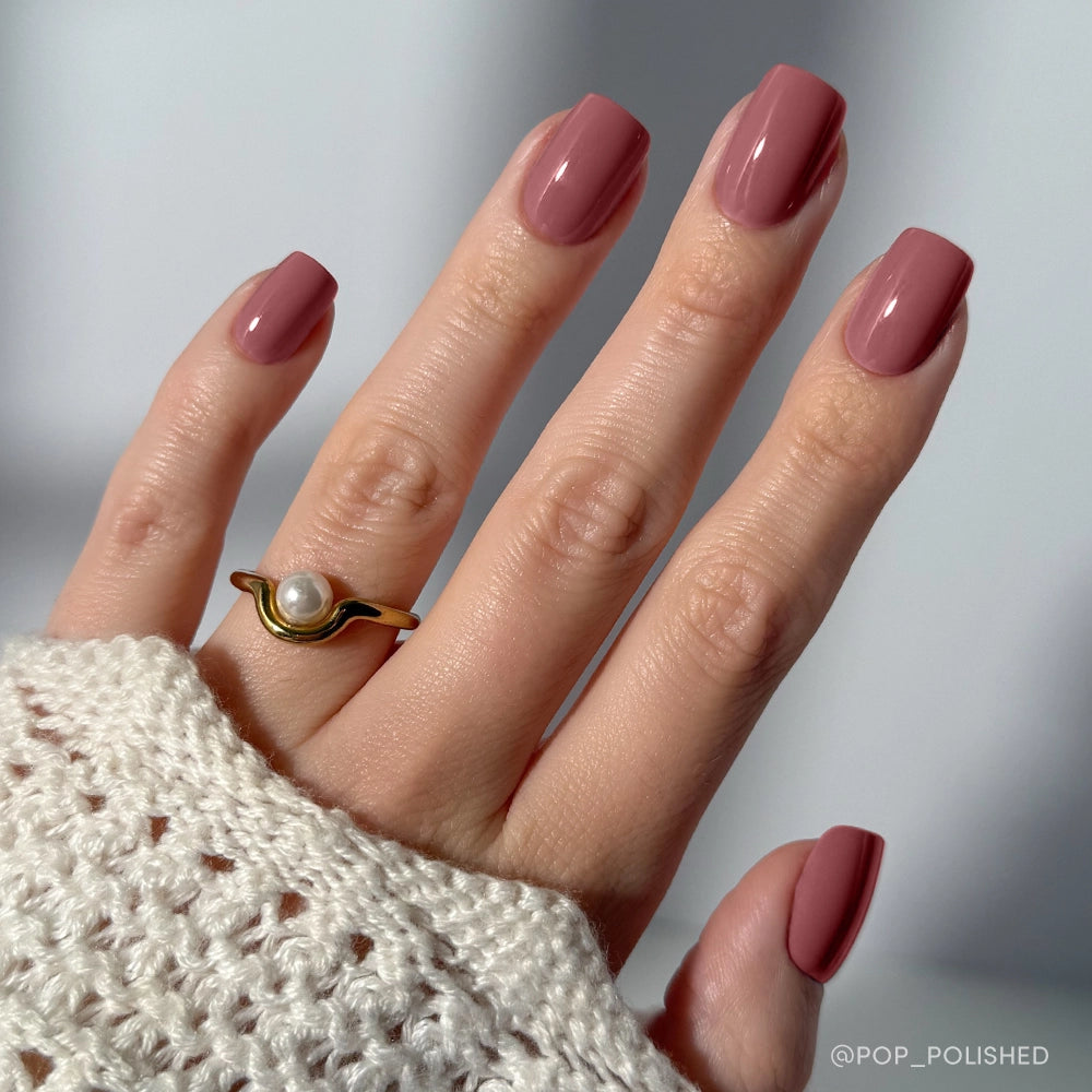 A hand with short squoval nails painted in glamnetic’s Mauve On. The person wears a gold pearl ring and a cream knit sweater. The background is softly blurred.