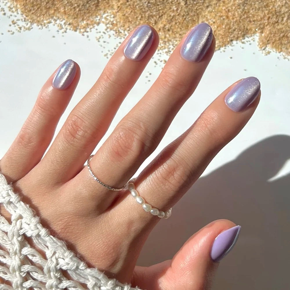 A hand adorned with glamnetic’s Mermaid’s Tail iridescent purple press-on nails, wearing delicate silver and pearl rings, rests on a surface sprinkled with grains and partially covered by a white crocheted sleeve.
