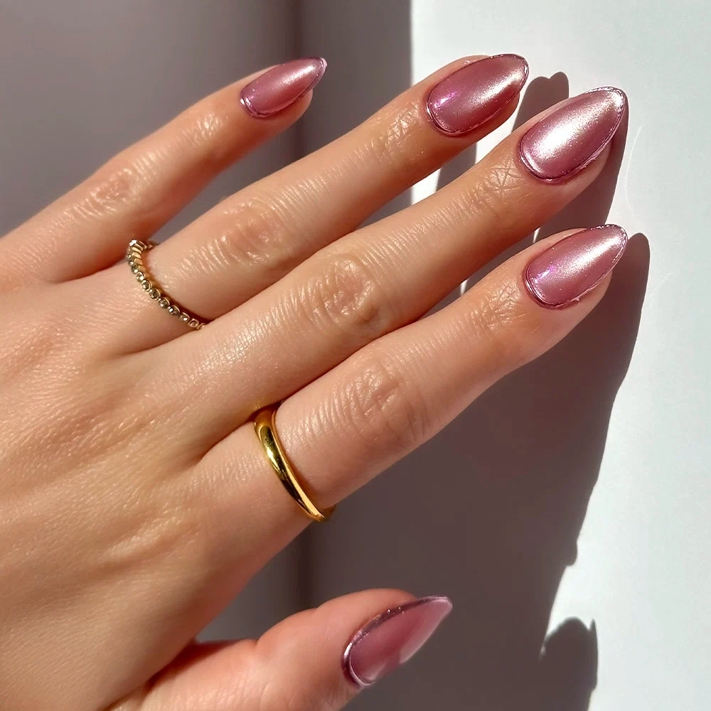 A hand with short, almond-shaped Glamnetic Mertini metallic pink press-on nails is shown against a white background. The hand wears a gold band ring and a delicate ring with small stones on the middle finger.