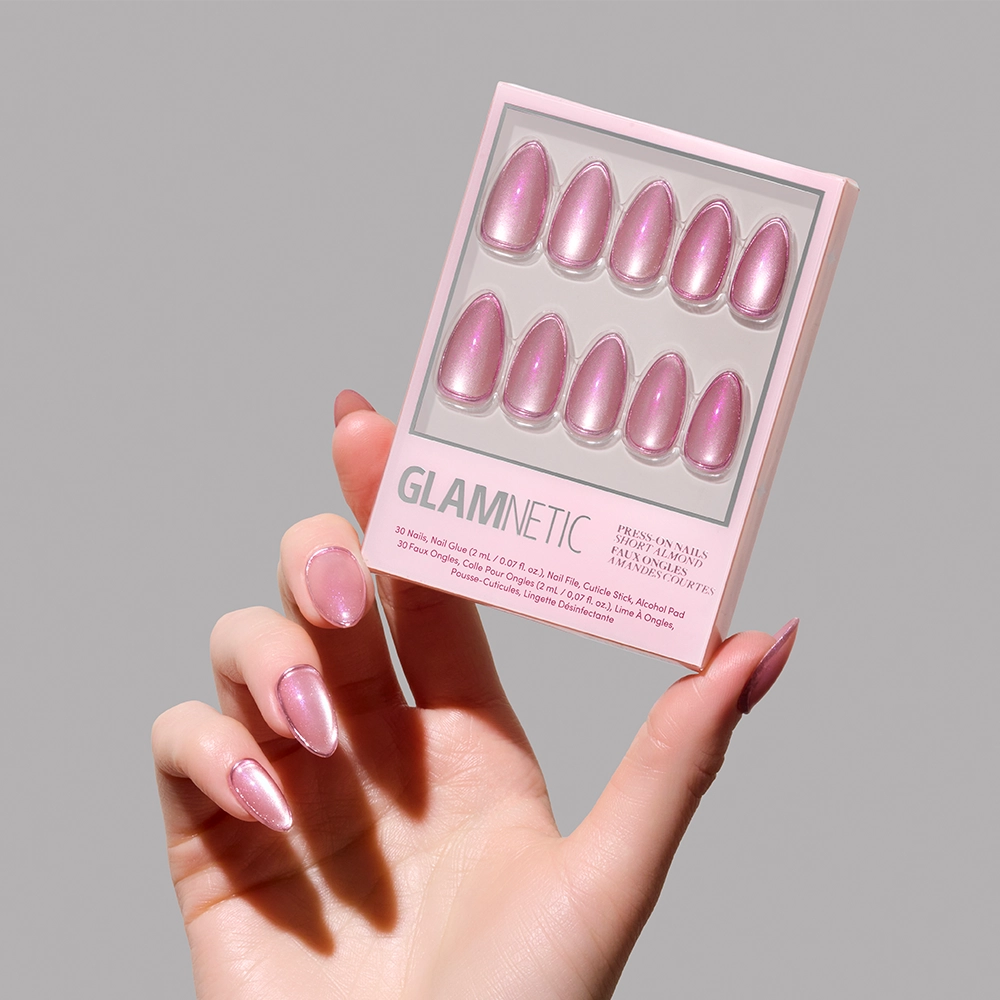 A hand with shimmering pink press-on nails holds the glamnetic Mertini Press-On Nail Set in a matching shade against a plain light gray background. The box displays 10 glossy, almond-shaped nails with a velvet finish in two rows. - Product image