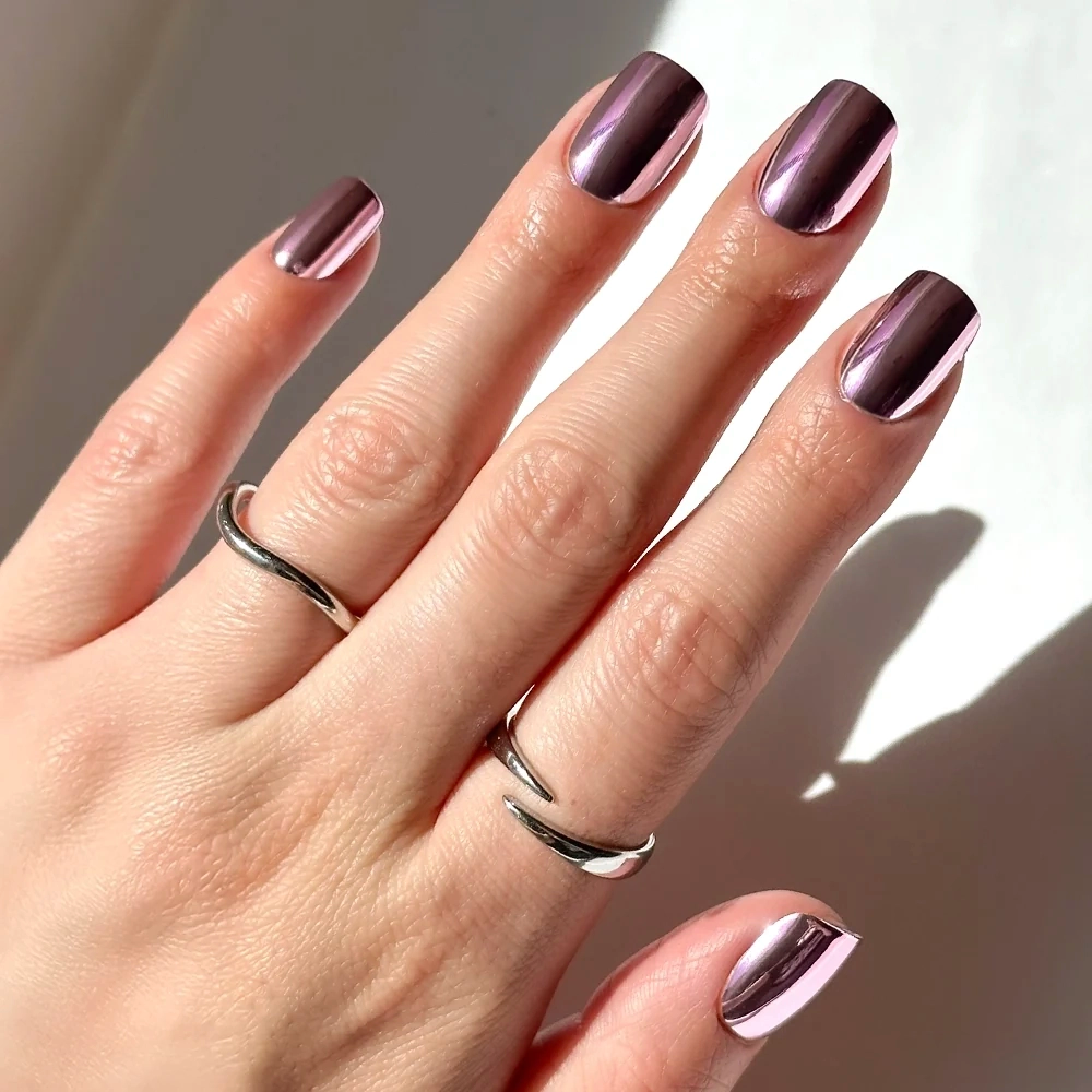 A hand with short squoval-shaped nails painted in glamnetic’s Metallic Blush, a pink metallic chrome finish, is shown against a light background, with two silver rings adorning the fingers.