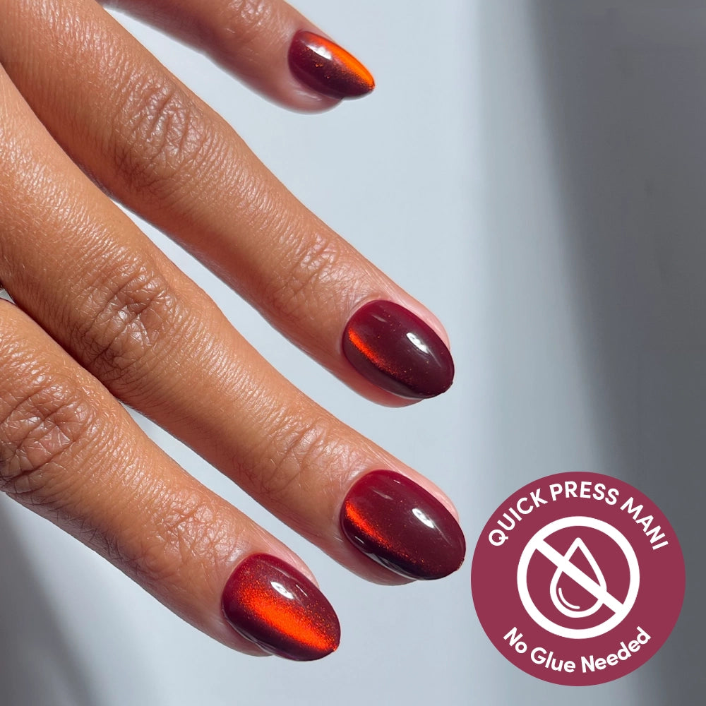 A hand displays glamnetic Midnight Merlot press-on nails in a shiny, deep red shade. A circular icon reads “Quick Press Mani, No Glue Needed,” emphasizing the effortless application of these stick-on nails. The background is light and neutral.
