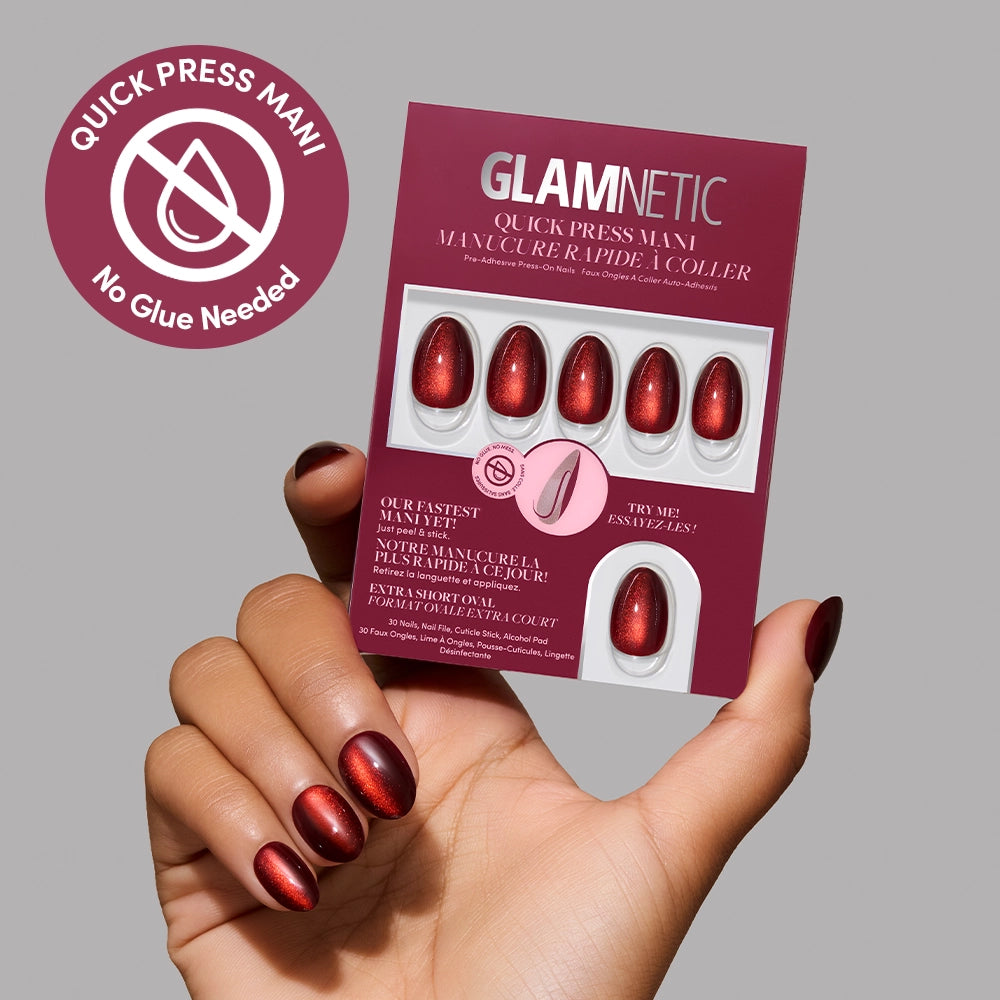 A hand holds a pack of glamnetic Midnight Merlot Quick Press Mani no-glue nails in shimmery red, with “No Glue Needed” text. These easy-to-use press-on nails are shown against a neutral background. - Product image