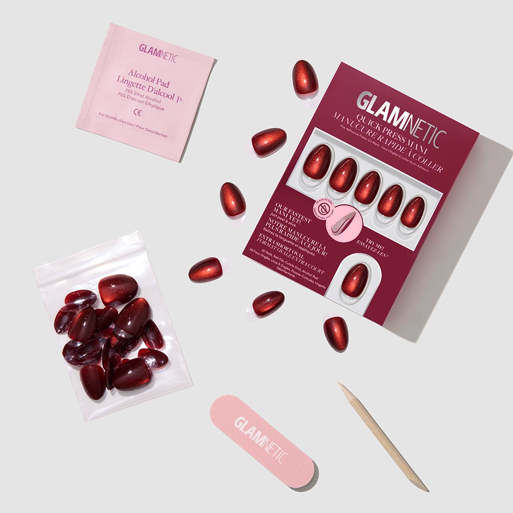Glamnetic Midnight Merlot press-on nail kit displayed on a light gray background. The set features a red box containing oval-shaped, dark red cat-eye stick-on nails, along with a bag of extra nails, a pink alcohol pad, a small nail file, and a wooden cuticle stick. - Product image