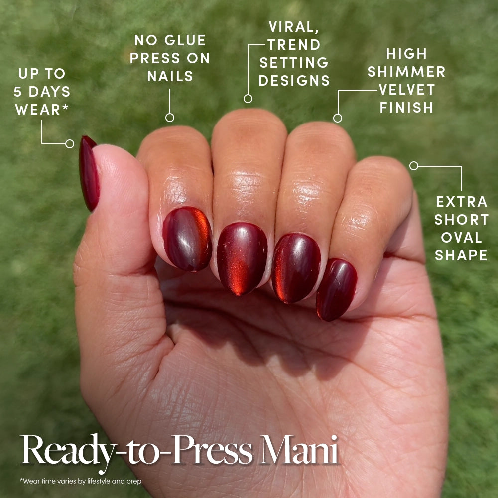 A hand with glossy, Midnight Merlot press-on nails by glamnetic in an extra short oval shape, held up outdoors. Text highlights up to 5 days wear, no glue needed with these convenient stick-on nails featuring a shimmer finish and trendy designs. - Product image