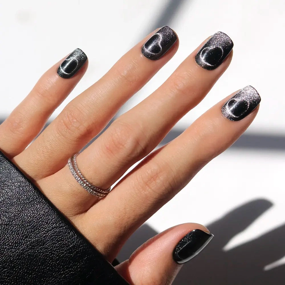 A hand with short squoval nails in glamnetic’s Midnight Romance shows off glossy black polish with abstract white designs, styled with a double-banded silver ring and black sleeve against a brightly lit background.
