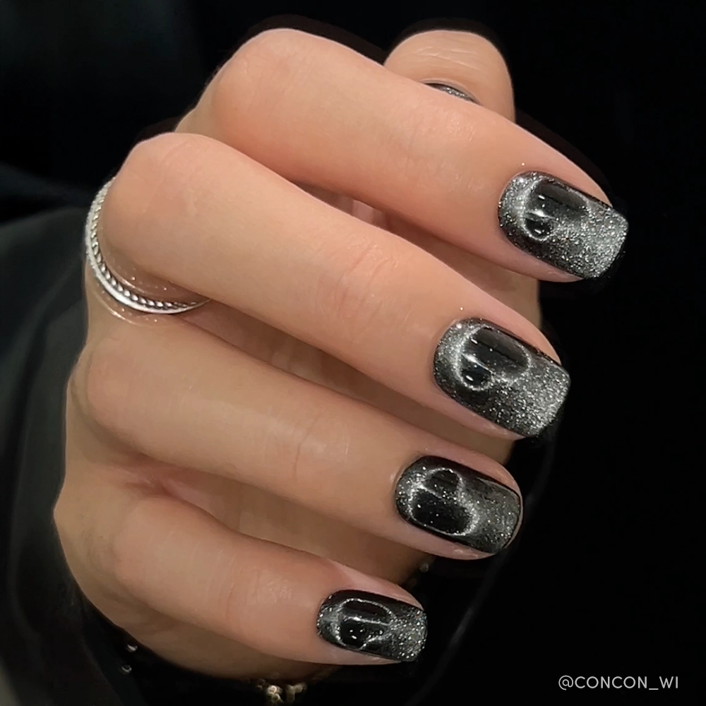 A hand with glamnetics Midnight Romance square nails featuring a black and silver glitter gradient, cat-eye effect, glossy finish, and clear droplet accents. The person also wears two silver rings. - Product image