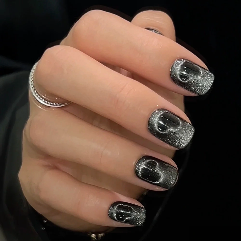 A hand with short squoval nails painted in glamnetic Midnight Romance—black with silver glitter and a shiny cat-eye effect—wears a silver ring on the middle finger. - Product image