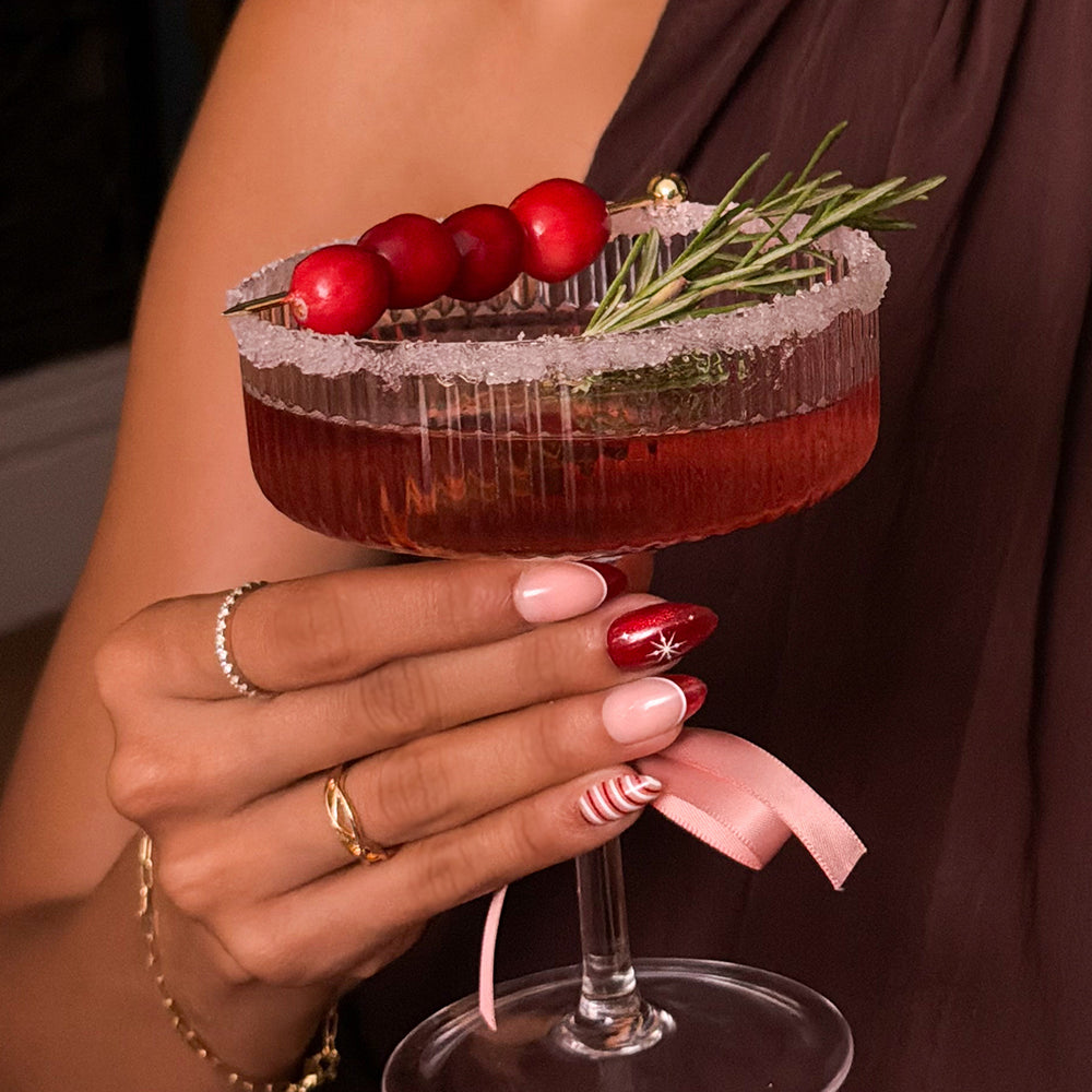 A hand wearing Glamnetic Mint to Be festive short almond nails with red French tips holds a red cocktail in a sugar-rimmed coupe glass garnished with cranberries and a sprig of rosemary. - Product image