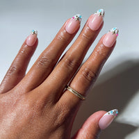 A hand adorned with a gold ring showcases manicured nails featuring a nude base and vibrant, jeweled white floral designs at the tips, reminiscent of the dazzling, jewel-encrusted look of glamnetic's Moonstone press-on nails.