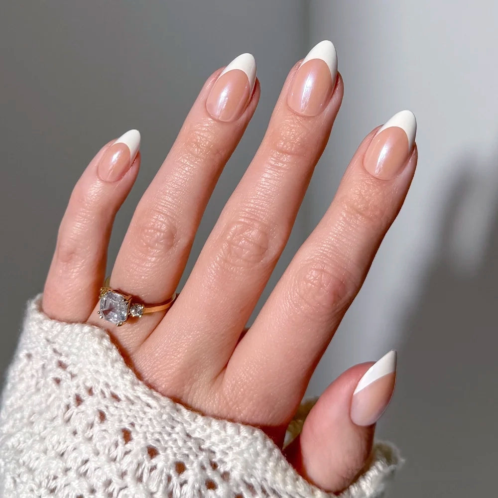 A hand with glamnetic’s “Mrs.” almond-shaped French tips is shown. The person wears a gold ring with a large gemstone, and a cream knit sweater sleeve partially covers the hand.