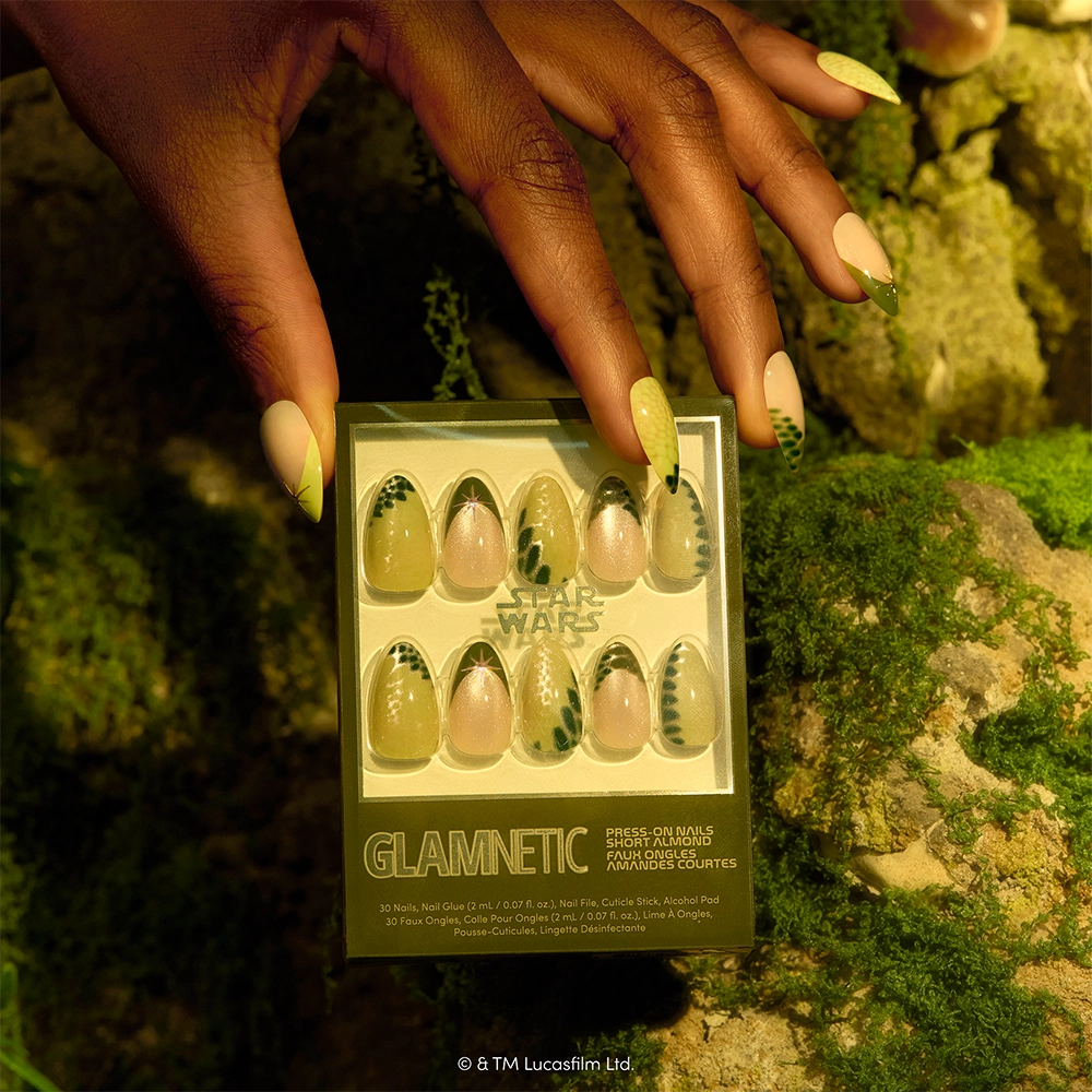 Much To Learn, You Still Have - A hand with green and black patterned nails holds a box of glamnetic Much To Learn, You Still Have press-on nails in a mossy, natural setting. The short almond nails feature matching green and black designs inspired by Yoda, giving them a unique galactic flair.