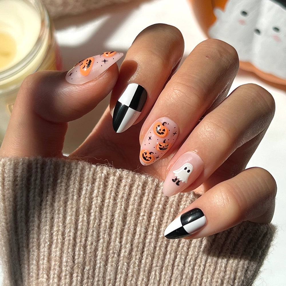 A hand with Halloween-themed nail art—featuring pumpkins, ghosts, and striking black-and-white geometric designs—rests on a beige knit sleeve, showcasing the short almond shape of glamnetic’s My Boo press-on nails, perfect for a spooky-season manicure.