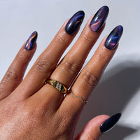 A hand with long, almond-shaped nails displays glamnetic's Mystic Topaz press-on nails, showcasing salon-quality, dark iridescent shades of purple and blue. Two gold rings—one geometric and one wavy—adorn the fingers against a plain light background, letting the enchanting finish of Mystic Topaz capture all attention.
