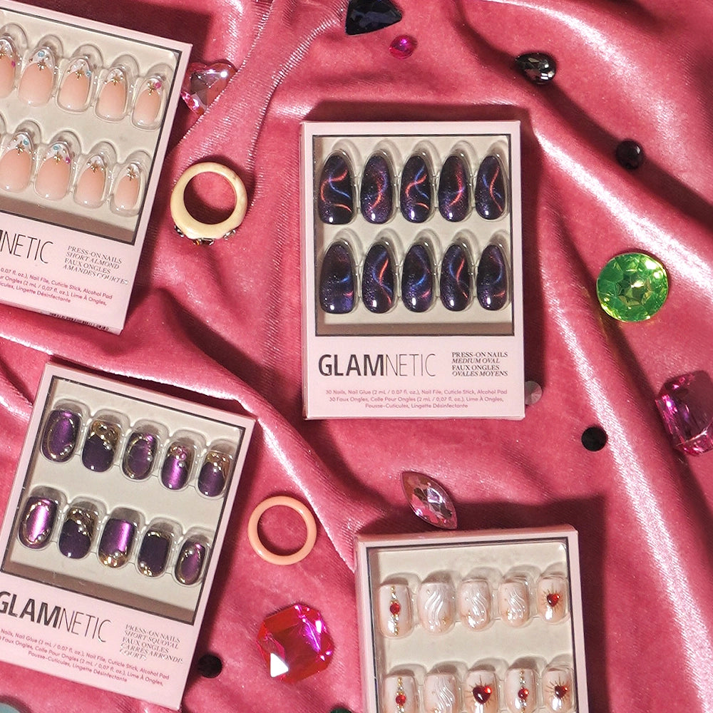Four glamnetic salon-quality press-on nail boxes in various colors and designs are displayed on pink satin fabric, surrounded by scattered gems, rings, and decorative crystals. Among them, the Mystic Topaz set stands out with its stunning iridescent finish, adding a touch of glamour to the luxurious scene. - Product image