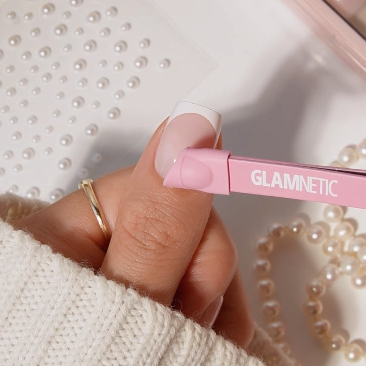 A hand with a gold ring and light pink French manicure uses glamnetic Nail Decal Tweezers to apply a pearl nail decoration, while pearl beads are scattered on a white textured surface in the background. - Product image