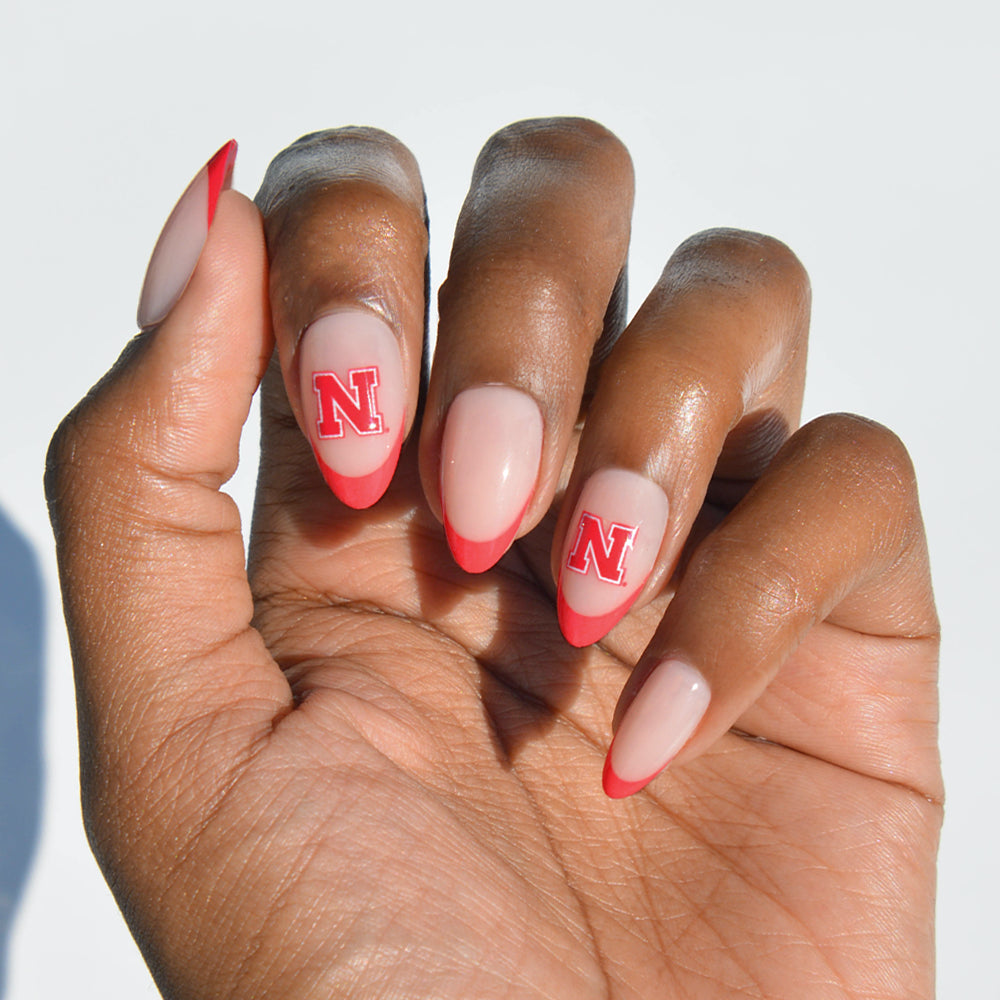 A hand with almond-shaped nails wearing glamnetic’s Collegiate Press-On Nails, featuring a nude base, red French tips, and red N logos on the middle and ring fingers. - Product image