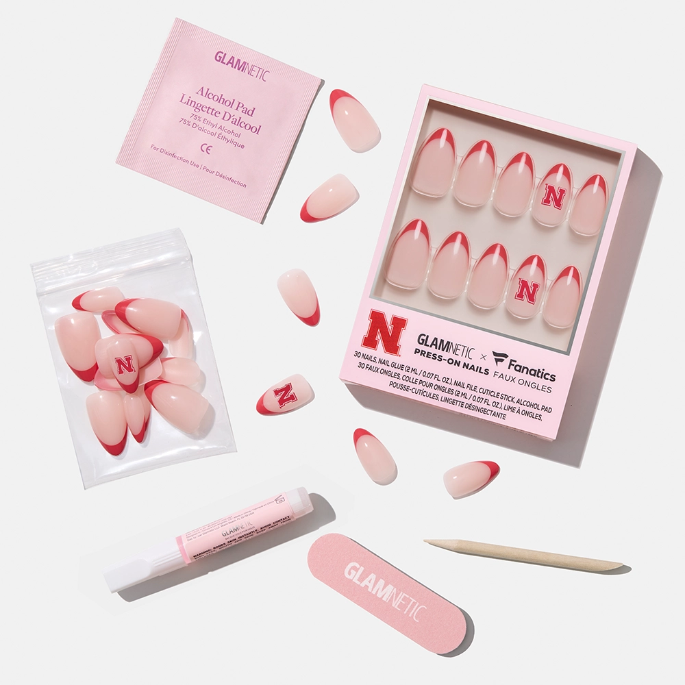 The glamnetic Nebraska Cornhuskers Press-On Nail Set, shown on a white background with a pink base, red tips, N logo nails, plus nail file, cuticle stick, alcohol pad, glue tube, and extra nails. - Product image