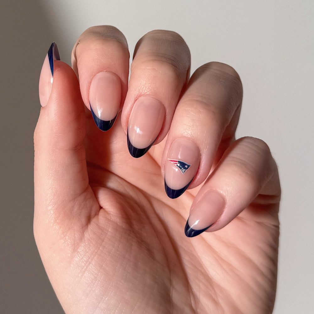 A hand showcases natural nails wearing glamnetic NFL Press-On Nails with dark blue French tips. One nail features a small New England Patriots logo as nail art. The background is plain and neutral. - Product image