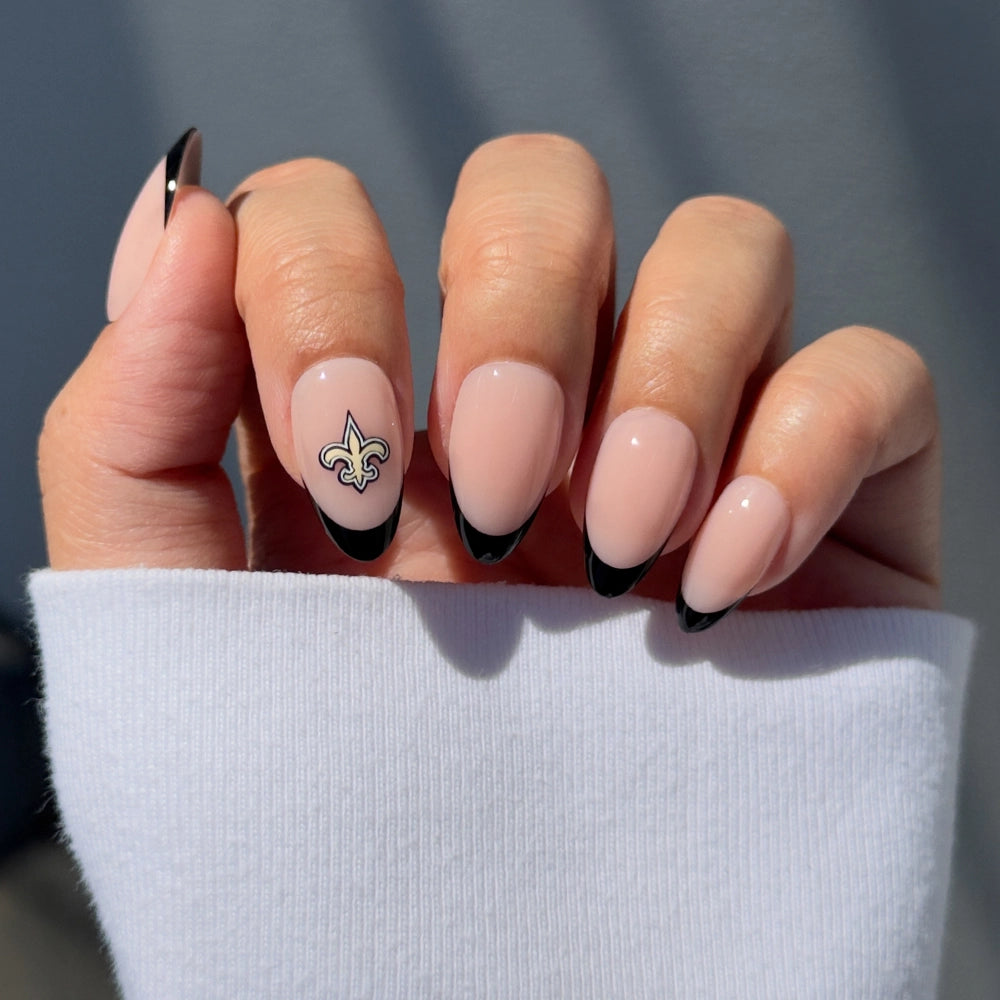 A hand wearing the glamnetic New Orleans Saints Press-On Nail Set features almond-shaped nails with a nude base, black French tips, and a gold fleur-de-lis on the ring finger. The person is dressed in a white long-sleeve top.
