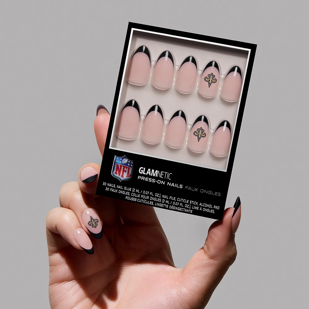 A hand displays the New Orleans Saints Press-On Nail Set by glamnetic, featuring black French tips and the team logo. The persons matching nails showcase this stylish, team-themed manicure.