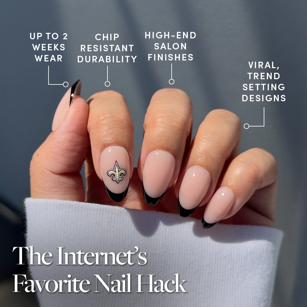 A hand shows off the glamnetic New Orleans Saints Press-On Nail Set with long, almond-shaped nails, a nude base, black French tips, and a gold fleur-de-lis. Overlay text highlights long wear, chip resistance, salon finish, and trendy designs. - Product image